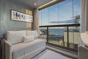 Varanda com vista para o mar, um sofá com manta estampada e um quadro abstrato emoldurado na parede.