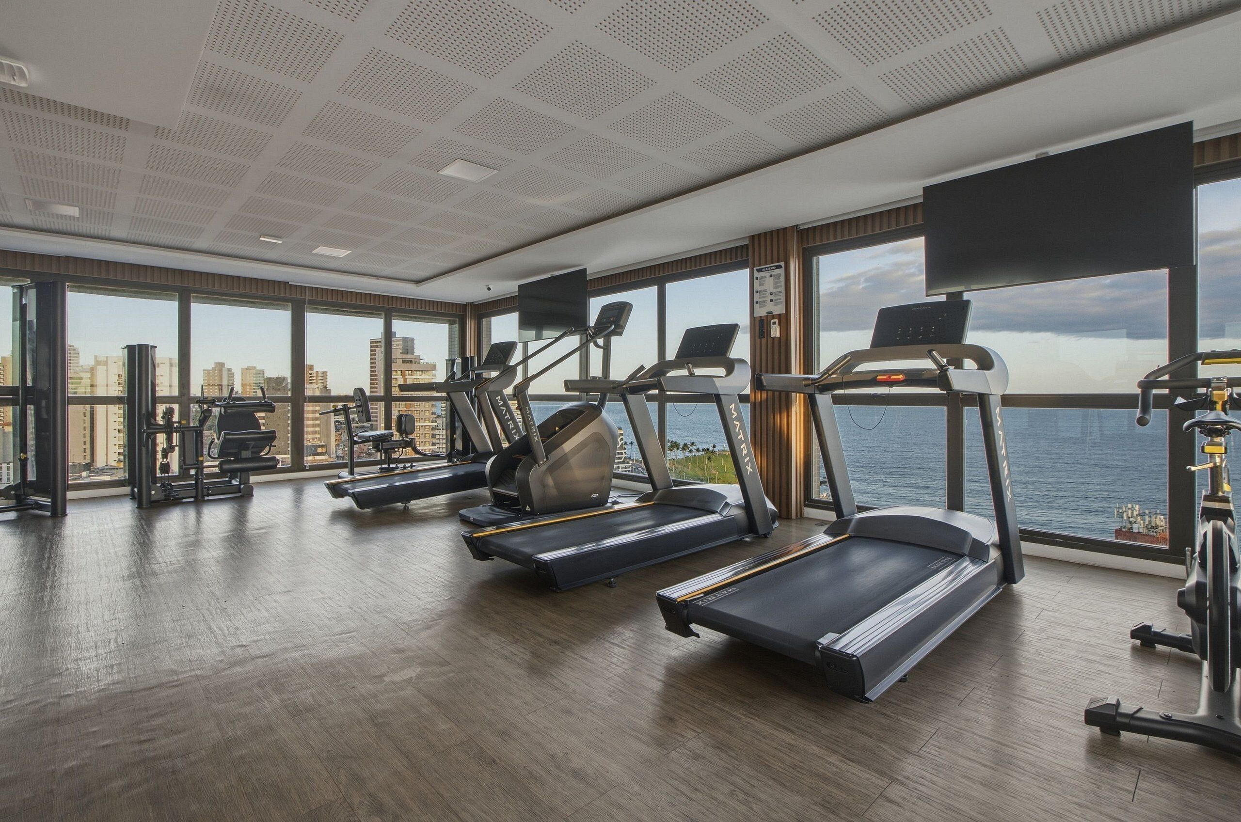 A gym with treadmills, elliptical machines, and weight equipment, large windows with a city view, and a high ceiling with a grid pattern.