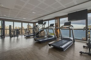 Academia com esteiras, elípticos e equipamentos de musculação, amplas janelas com vista para a cidade e teto alto com padrão de grade.
