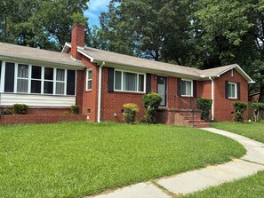 A two-story brick house with a front porch, a chimney, and a well-maintained lawn. - West side gem on a half-acre lot. Sunroom for indoor/outdoor living. Book today! (Charlotte)