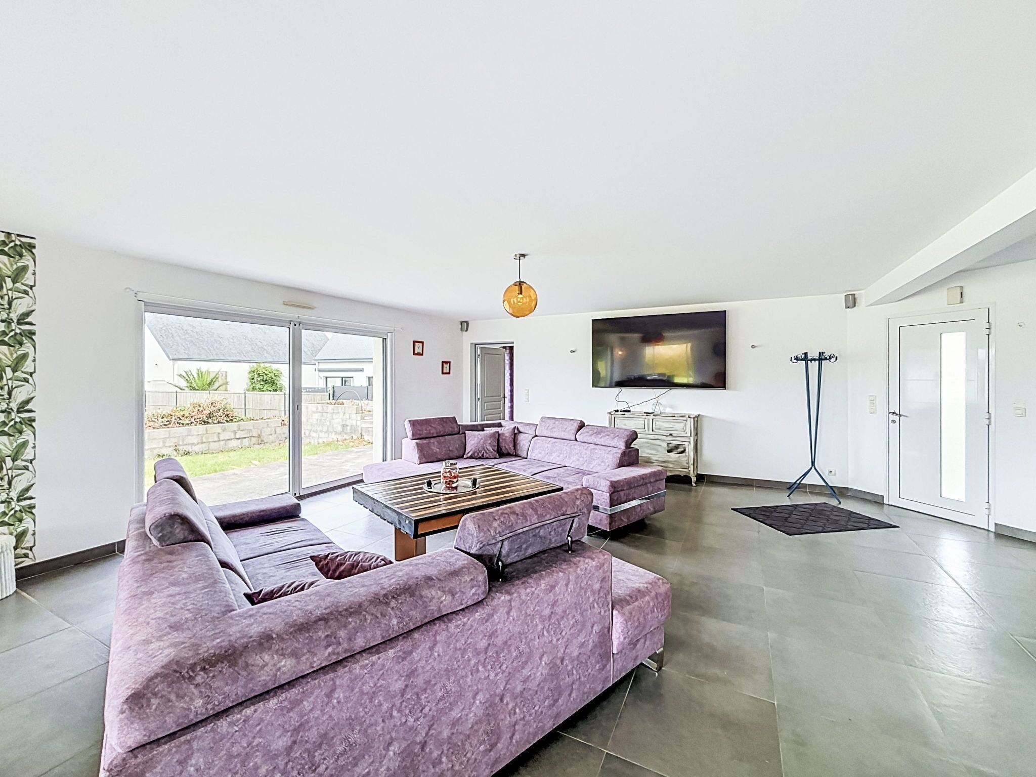 A modern living room with a sectional sofa, a coffee table, a TV mounted on the wall, and a sliding glass door leading to an outdoor area.