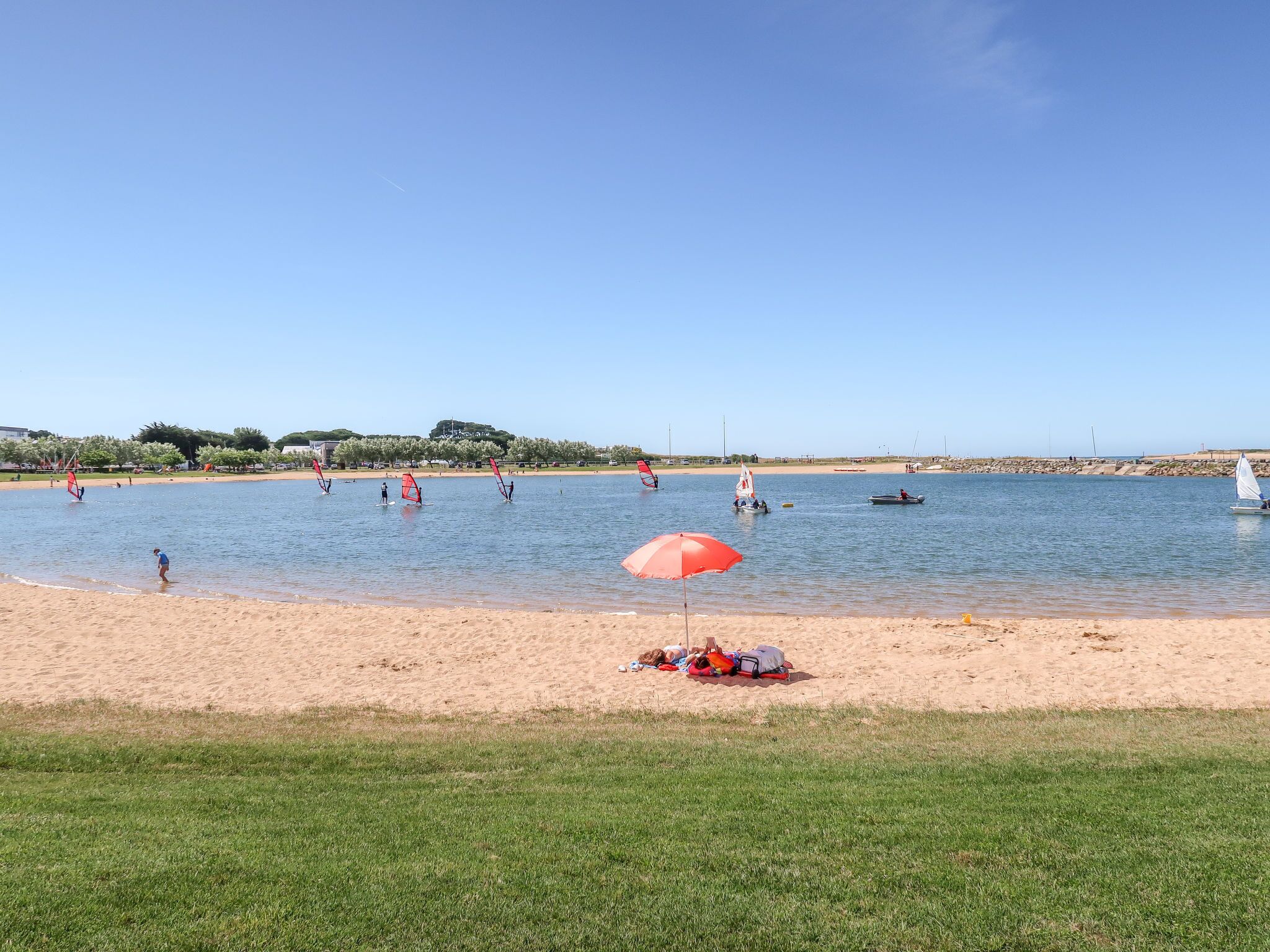 Ein Strand mit Windsurfenden, einer Segelyacht und einem roten Sonnenschirm im Sand.
