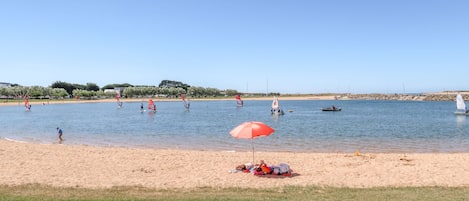 Ein Strand mit Windsurfenden, einer Segelyacht und einem roten Sonnenschirm im Sand.