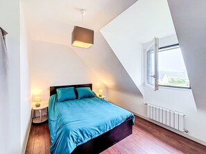Ein Schlafzimmer mit schrägen Wänden, einem Bett mit blauer Bettwäsche, zwei Nachttischlampen, einem Fenster mit Aussicht und einer Heizung.