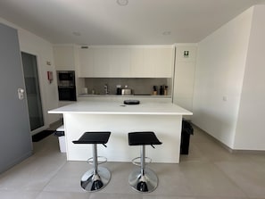 A modern kitchen with white cabinets, a central island with stools, a built-in oven, and a fire extinguisher. - Home Sweet Home. (Parchal)