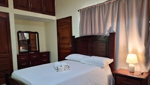 A neatly made bed with a white comforter and pillows, a bedside table with a lamp, wooden furniture, and a mirror on the wall. - Estudio With Bed, Tv, Ac, Kitchen, Refri and Bacony. (Santo Domingo)