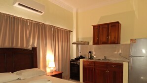 A hotel room with a bed, a nightstand with a lamp, a kitchenette with a stove, and wooden cabinets. - Estudio With Bed, Tv, Ac, Kitchen, Refri and Bacony. (Santo Domingo)