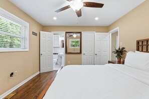 Une chambre à coucher comprenant un lit, un ventilateur de plafond, une fenêtre avec des stores et une porte donnant sur une salle de bain.