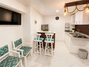 Une cuisine compacte avec des meubles blancs, une cuisinière intégrée et un petit coin repas avec des chaises blanches et des coussins turquoise.