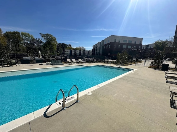 A rectangular swimming pool with clear blue water, surrounded by lounge chairs and a concrete deck.