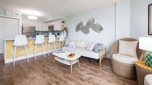 Een moderne keuken met witte kasten, een roestvrijstalen koelkast en een magnetron. Er is een eethoek met een witte tafel en stoelen, plus een woonkamer met een bank en een rieten fauteuil.
