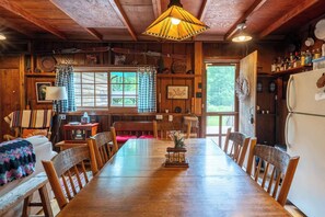 A wooden dining room with a long table, chairs, a lamp, and a refrigerator.