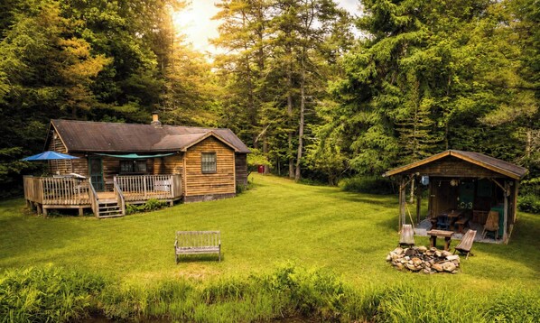 A rustic cabin with a deck and a small porch, surrounded by a lush green lawn and dense forest.
