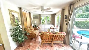 Ein sonnendurchfluteter Raum mit Deckenventilator, einem großen Fenster mit Vorhängen, einer Couch, einem Korbsessel und einem Blumentopf.