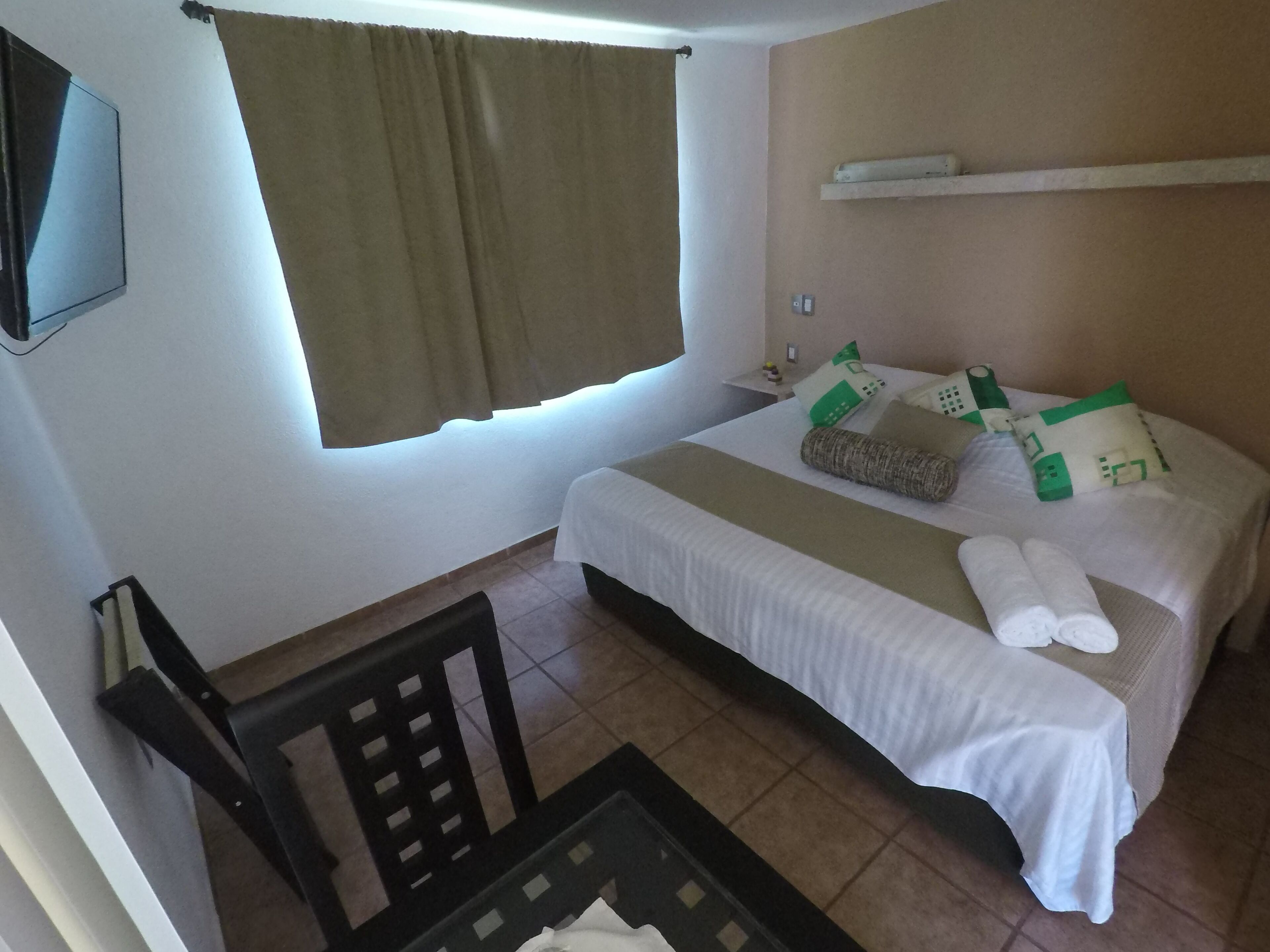 Una habitación de hotel con cama, sillón, televisión y ventana con cortinas.