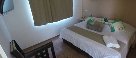 Una habitación de hotel con cama, sillón, televisión y ventana con cortinas.