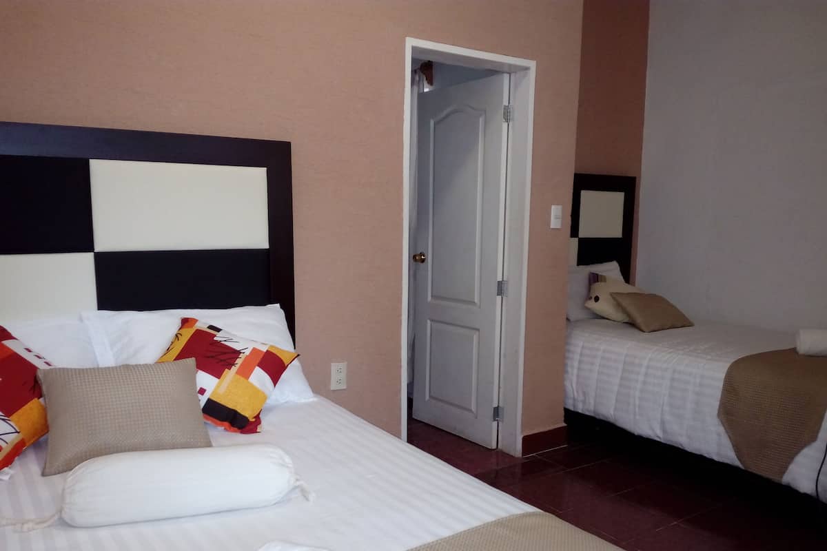 A hotel room with two beds, one with a headboard and the other with a plain headboard. There is a door between the beds, and a mirror on the wall.