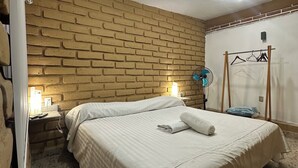 Un dormitorio con pared de ladrillo, una cama con cubrecama blanco, una mesita de noche con lámpara y un ventilador de techo.