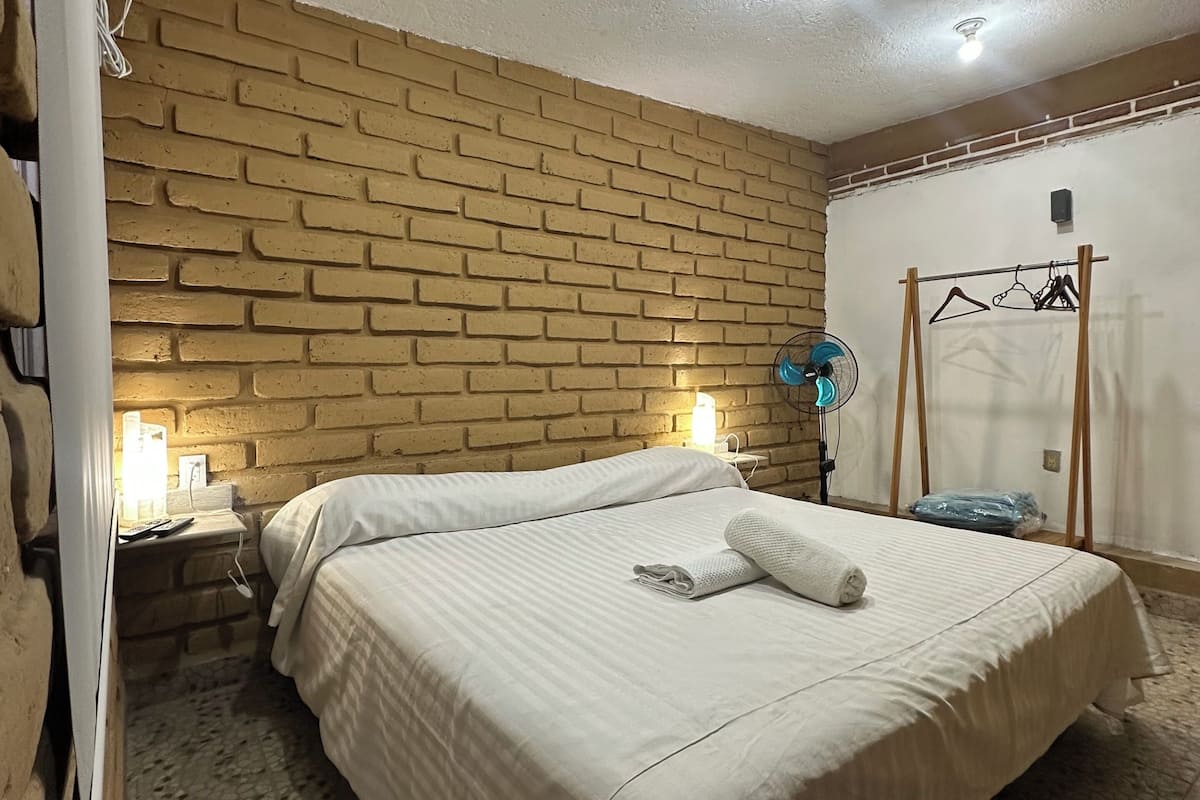 A bedroom with a brick-patterned wall, a bed with a white bedspread, a bedside table with a lamp, and a ceiling fan.