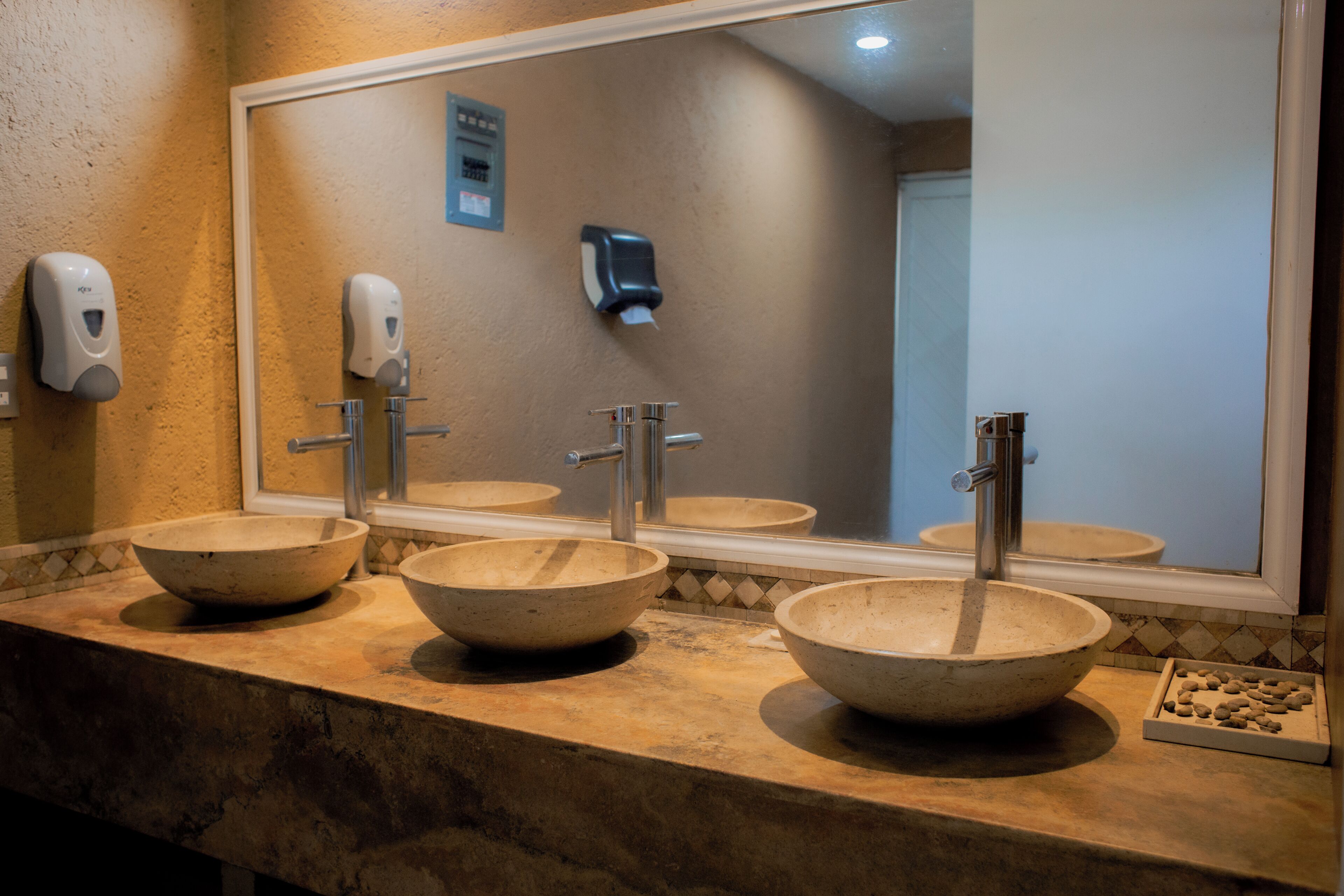 Un baño con tres lavamanos grandes, redondos y color beige, un espejo y un dispensador de jabón en la pared.
