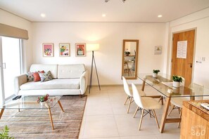 A modern living room with a sofa, coffee table, dining table, and chairs. There are framed pictures on the wall, a floor lamp, and a mirror.