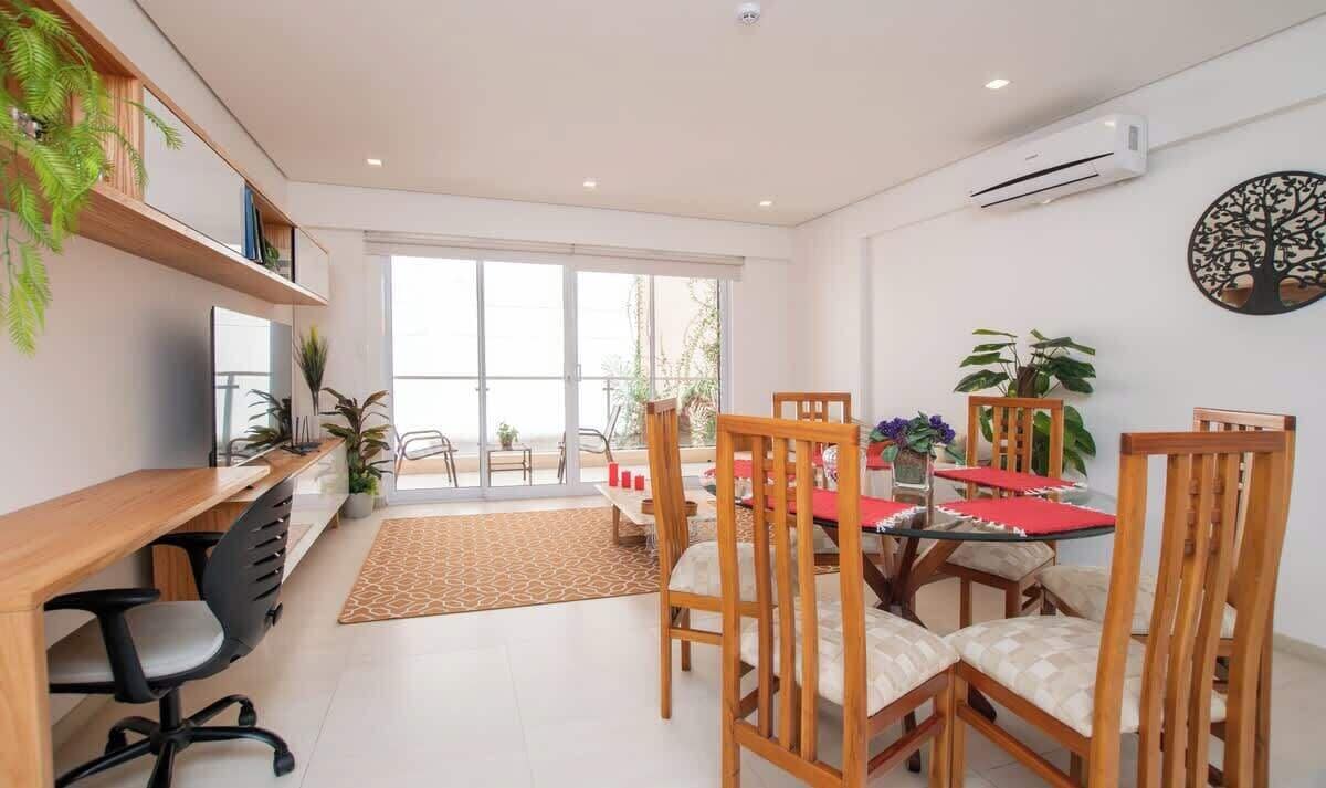 A modern dining area with a wooden table, chairs, and a red table runner. There are potted plants and a large window with a view.