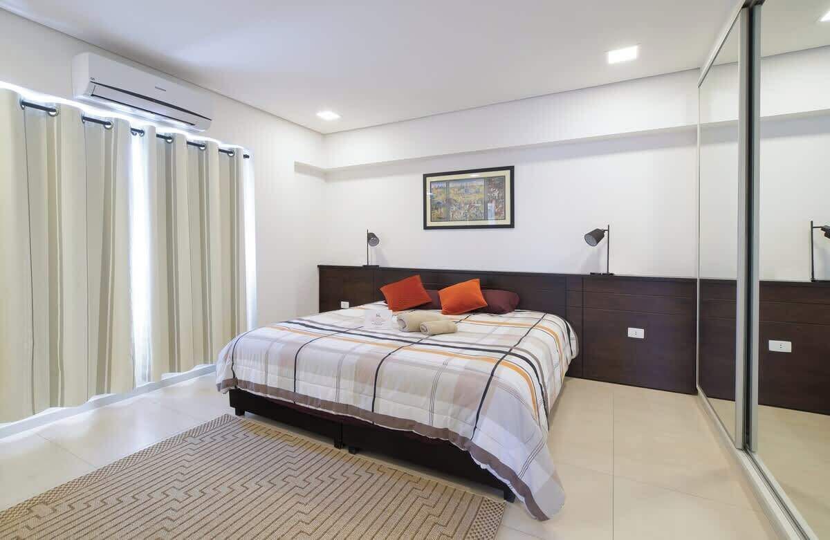 A bedroom with a bed, a window with curtains, an air conditioning unit, a framed picture on the wall, and a bedside lamp.