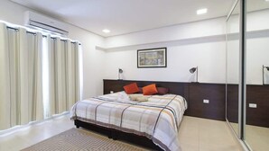 Un dormitorio con cama, ventana con cortinas, equipo de aire acondicionado, un cuadro en la pared y una lámpara de noche.