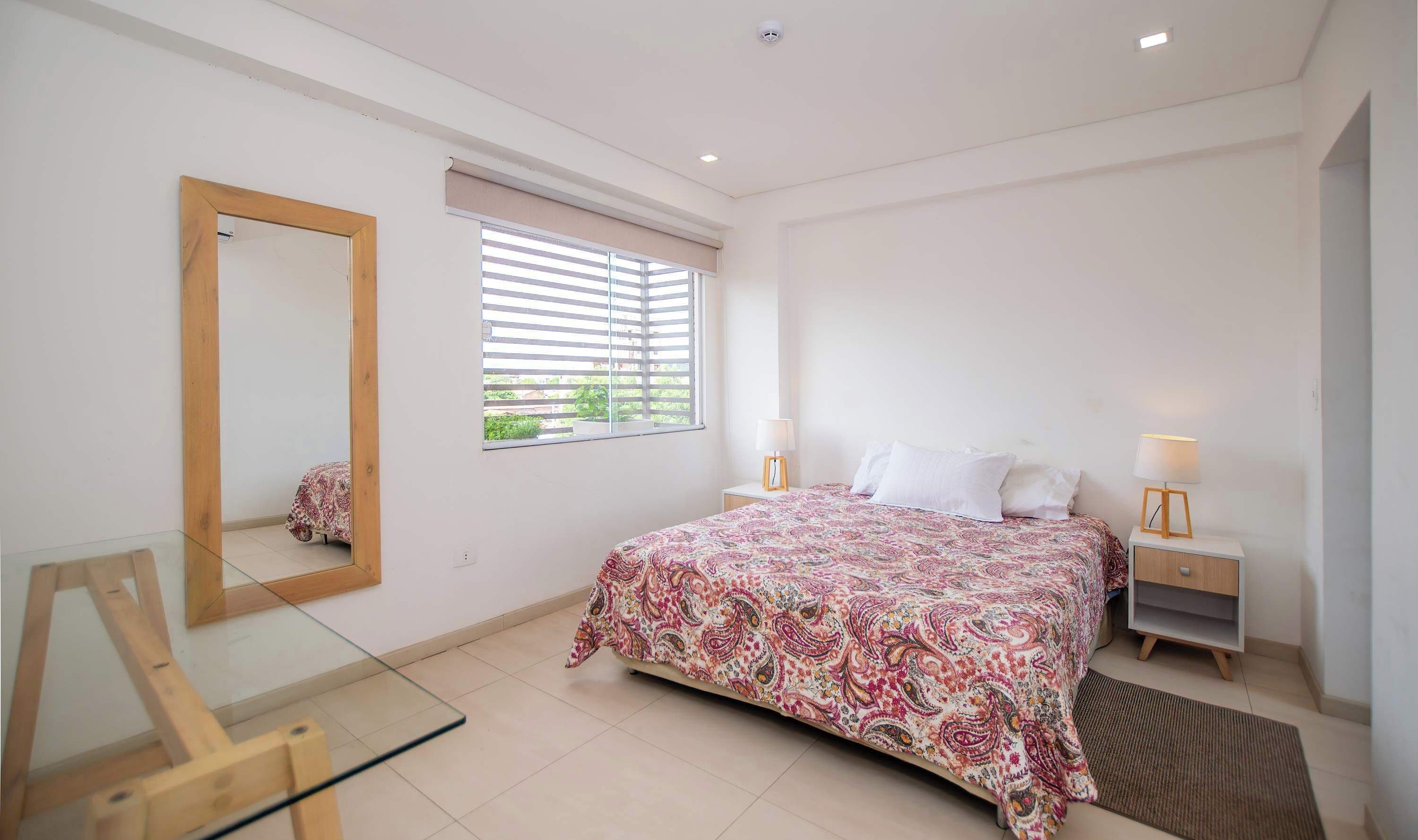 A bedroom with a bed, bedside tables, a mirror, a window with blinds, and a glass table.