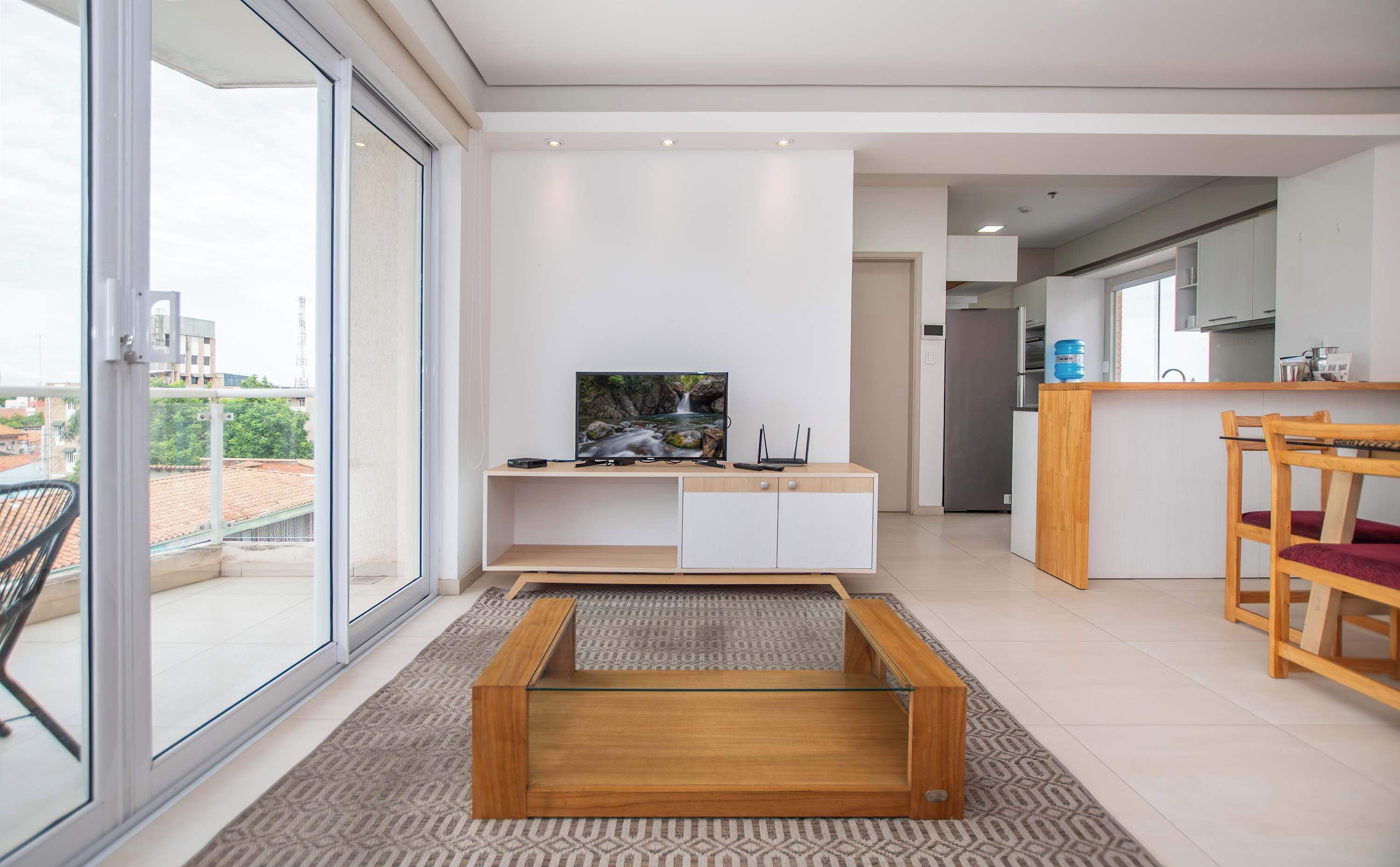 A modern living room with a TV, a glass coffee table, and a kitchen area in the background.