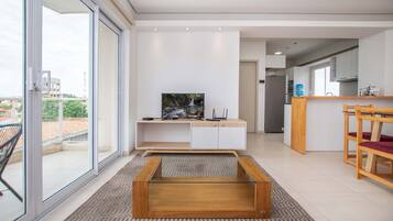 A modern living room with a TV, a glass coffee table, and a kitchen area in the background.