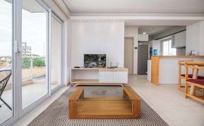 Una sala de estar moderna con televisor, una mesa de centro de vidrio y una cocina al fondo.