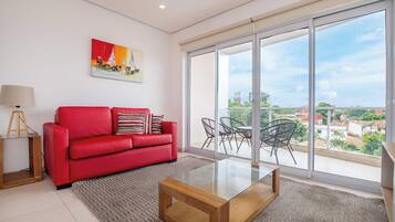 Una sala de estar moderna con un sofá rojo, una mesa de centro de vidrio, un cuadro y un amplio ventanal corredera que da a un balcón exterior con vistas a edificios y zonas verdes.