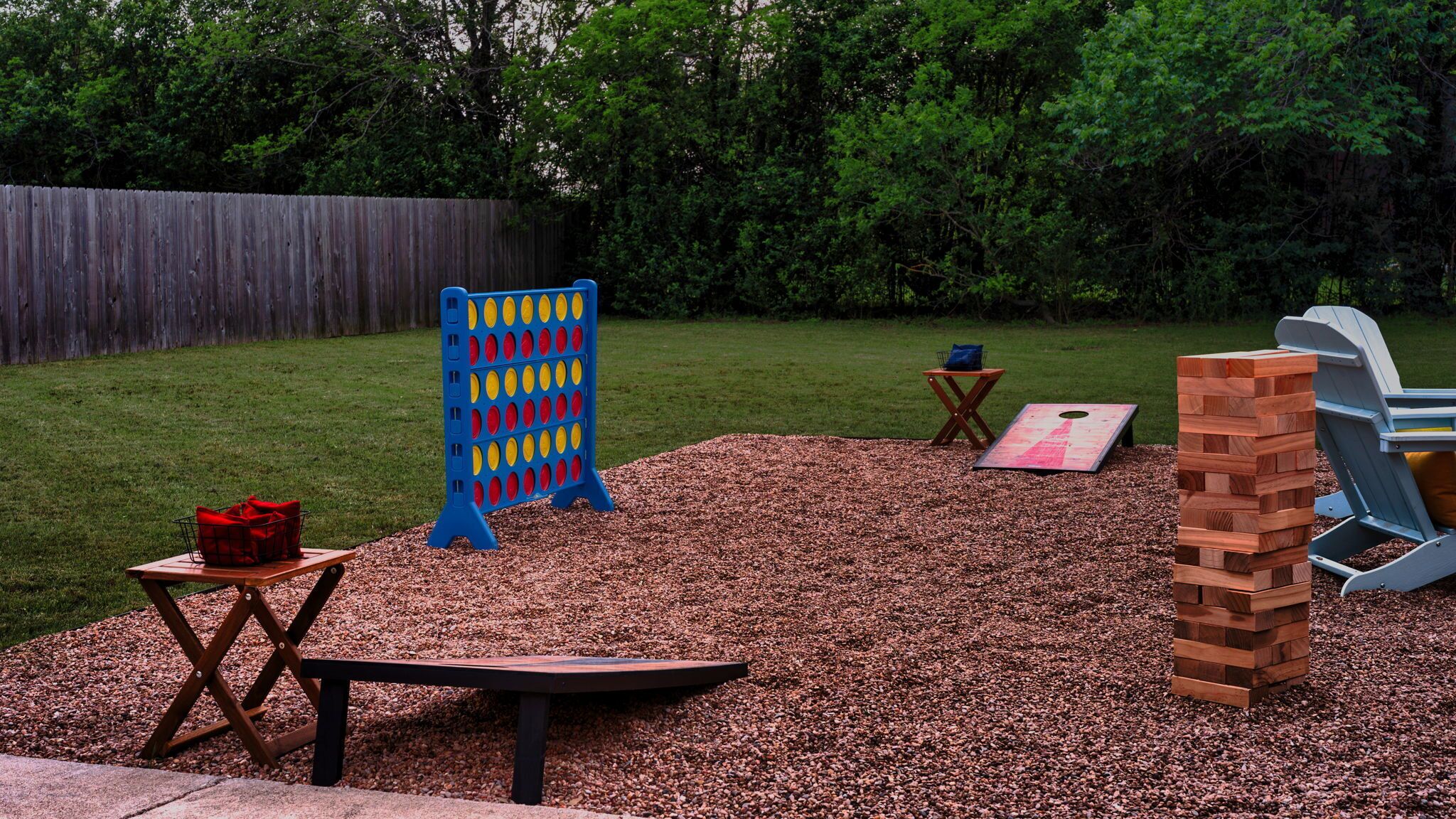 Un patio trasero con cerca de madera, un tablero de juego azul y rojo, una mesa de madera, una silla azul y una estructura de madera.