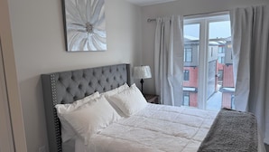 Un dormitorio con un cabecero capitonado gris, ropa de cama blanca, un adorno en la pared, una lámpara de noche y una ventana con cortinas.