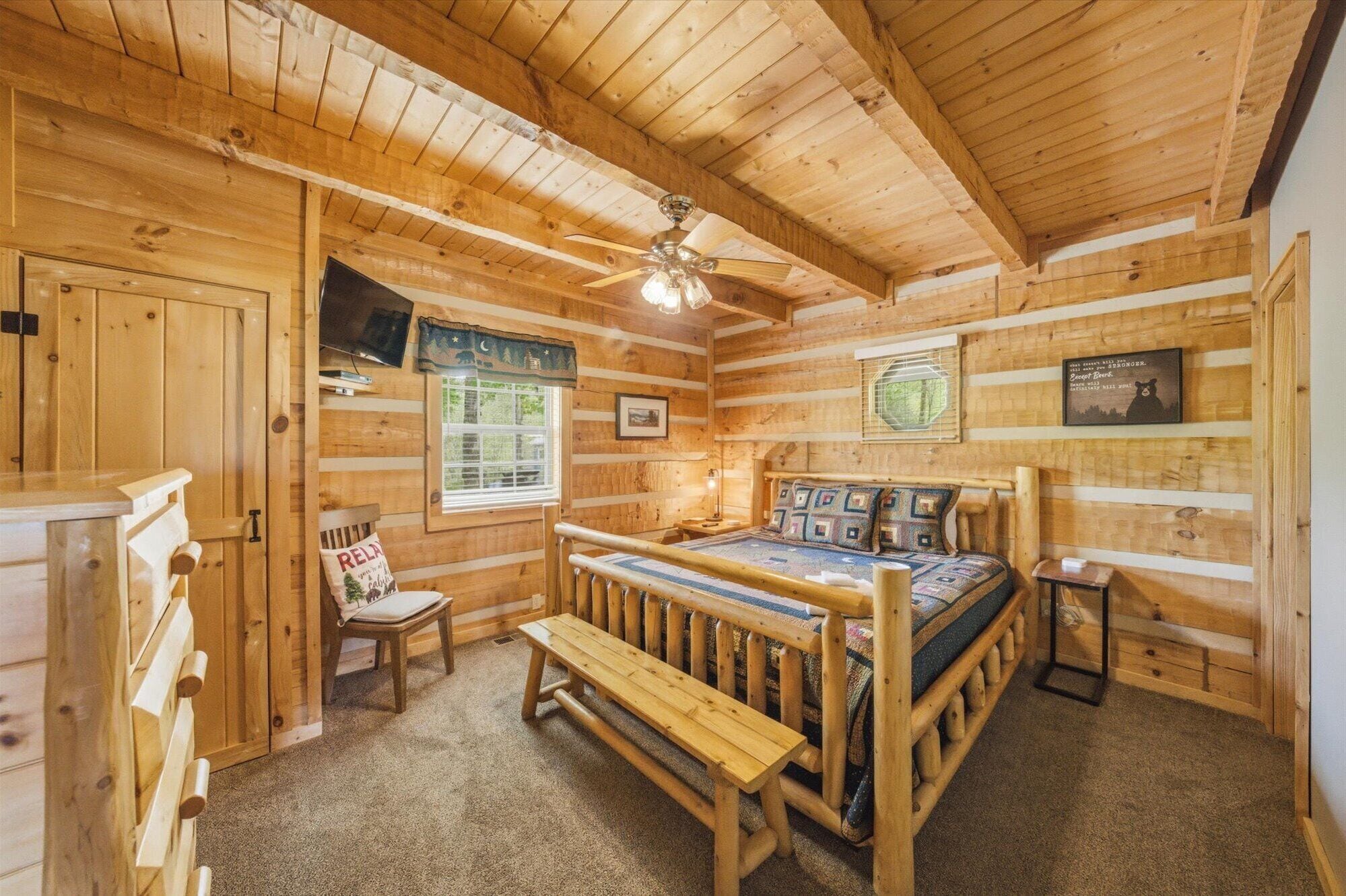 Un dormitorio tipo cabaña de troncos con una cama de madera, un banco, un ventilador de techo y una ventana con vistas a los árboles.