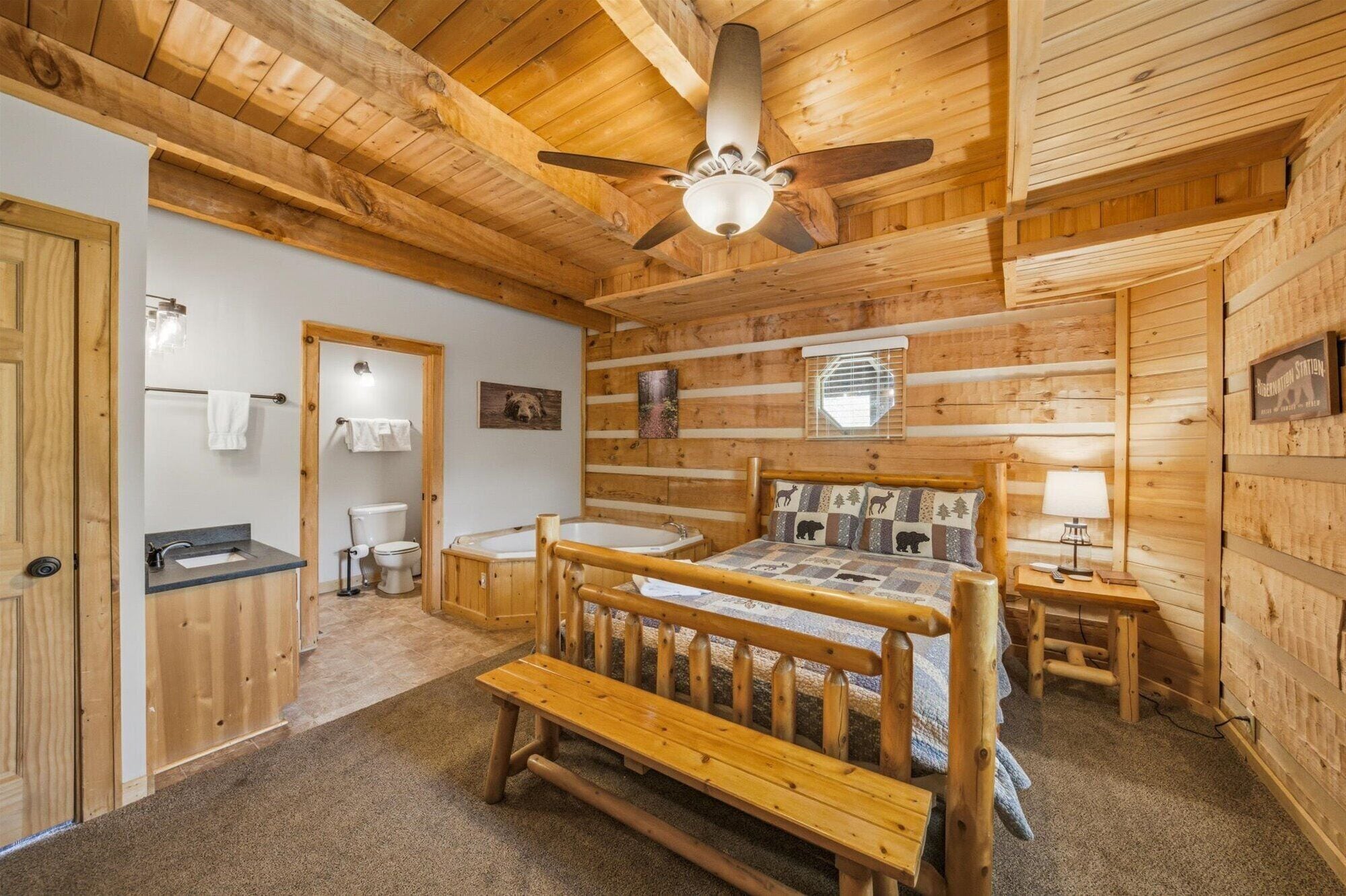 A log cabin bedroom with a wooden bed, a ceiling fan, a bathroom with a toilet and sink, and a small table with a lamp.