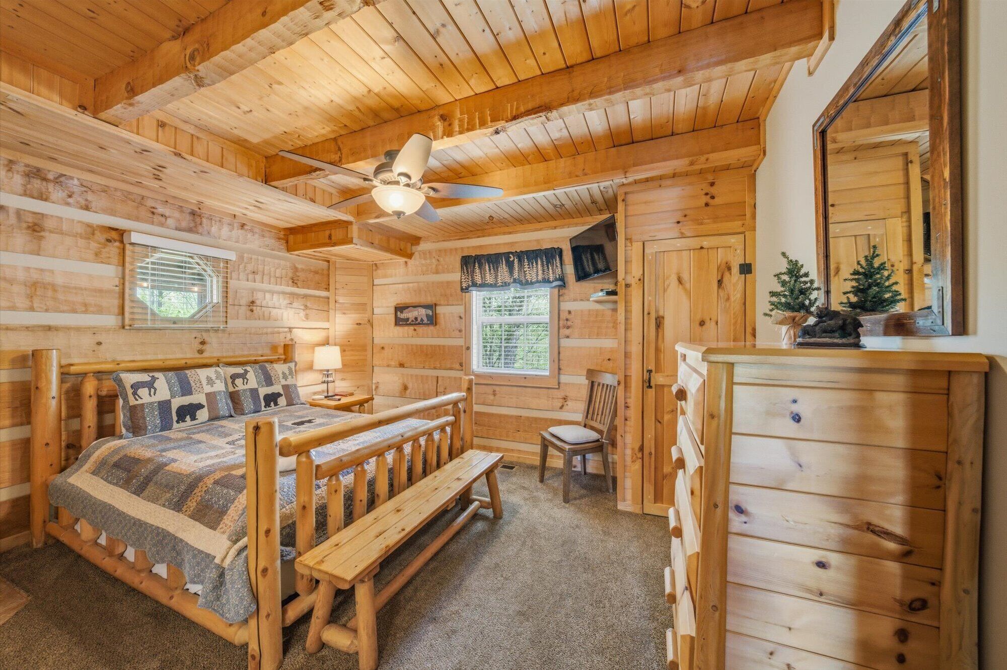 A log cabin room with a bed, a desk, a chair, a dresser, and a ceiling fan.