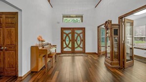 A spacious room with wooden flooring, a piano, a large wooden door with glass panels, and a view through a glass door. - Blue Oasis (College Station)