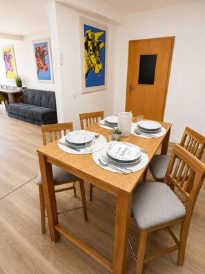 A dining area with a wooden table set for four, surrounded by chairs, in a room with a sofa and artwork on the walls. - Nice Flats Premium 4-Room, 6-Bed Apartment, 3 Bedrooms, Smart TV, Kitchen (Hamm)