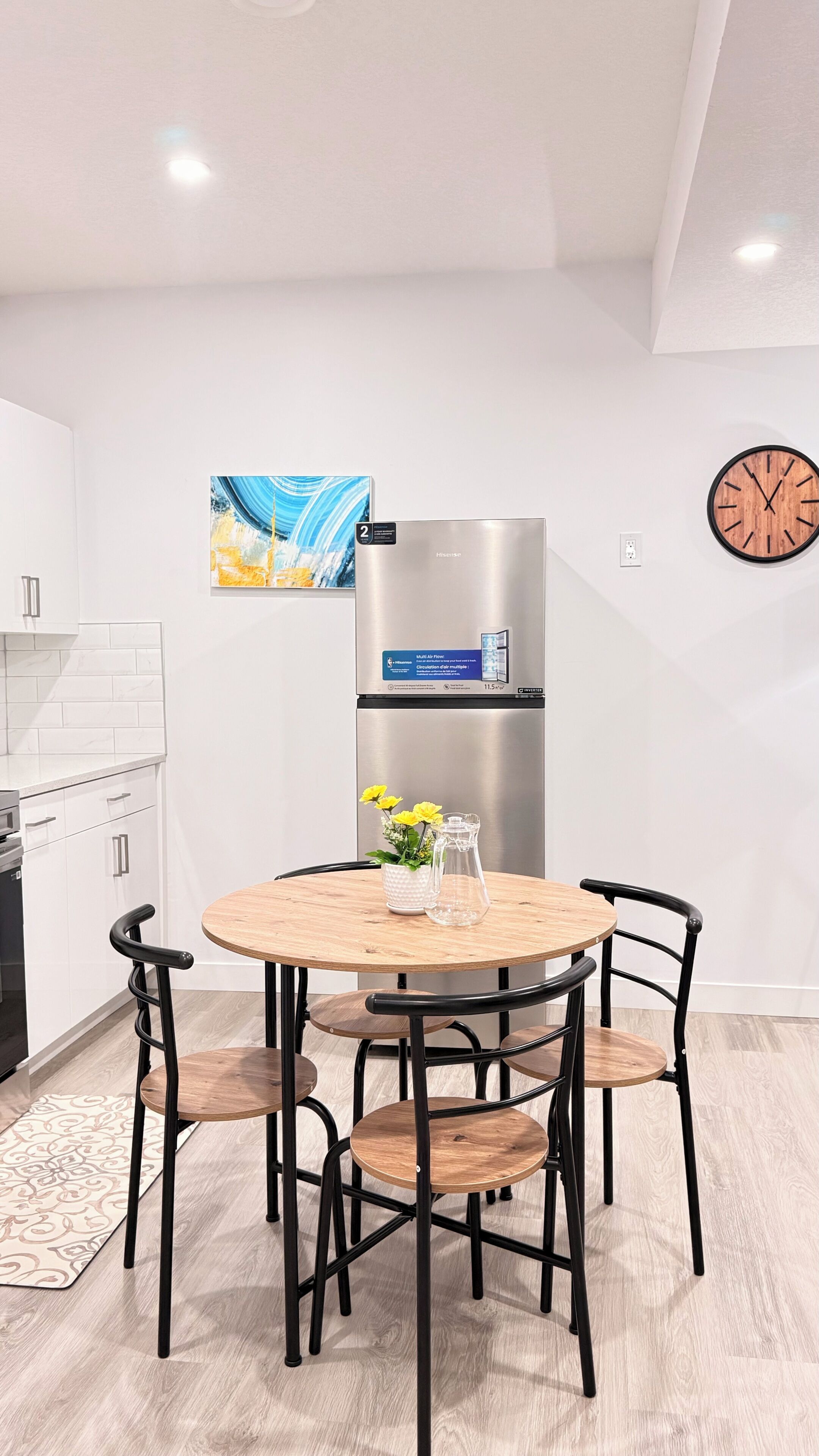 A modern kitchen with white cabinetry, a stainless steel refrigerator, a round wooden dining table with four chairs, and a wall-mounted clock.