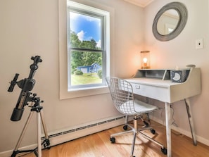 A room with a desk, chair, telescope, and a window with a view of a blue house.