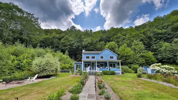 Ein blaues, zweistöckiges Haus mit Veranda, umgeben von üppiger Vegetation und blauem Himmel.