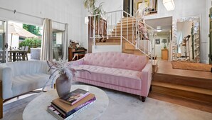 Un saló modern amb un sofà rosa, un butac armchair blanc, una taula de centre rodona amb llibres, una escala i un mirall gran.