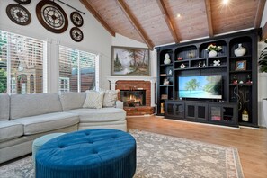 A living room with a fireplace, a large TV, a sofa, and a blue ottoman. - Main House Santa Clarita Retreat+Pets with fee (Santa Clarita)