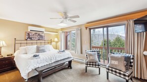 Un dormitorio con cama, lámparas de noche, ventilador de techo, ventana grande con cortinas y vista a árboles.