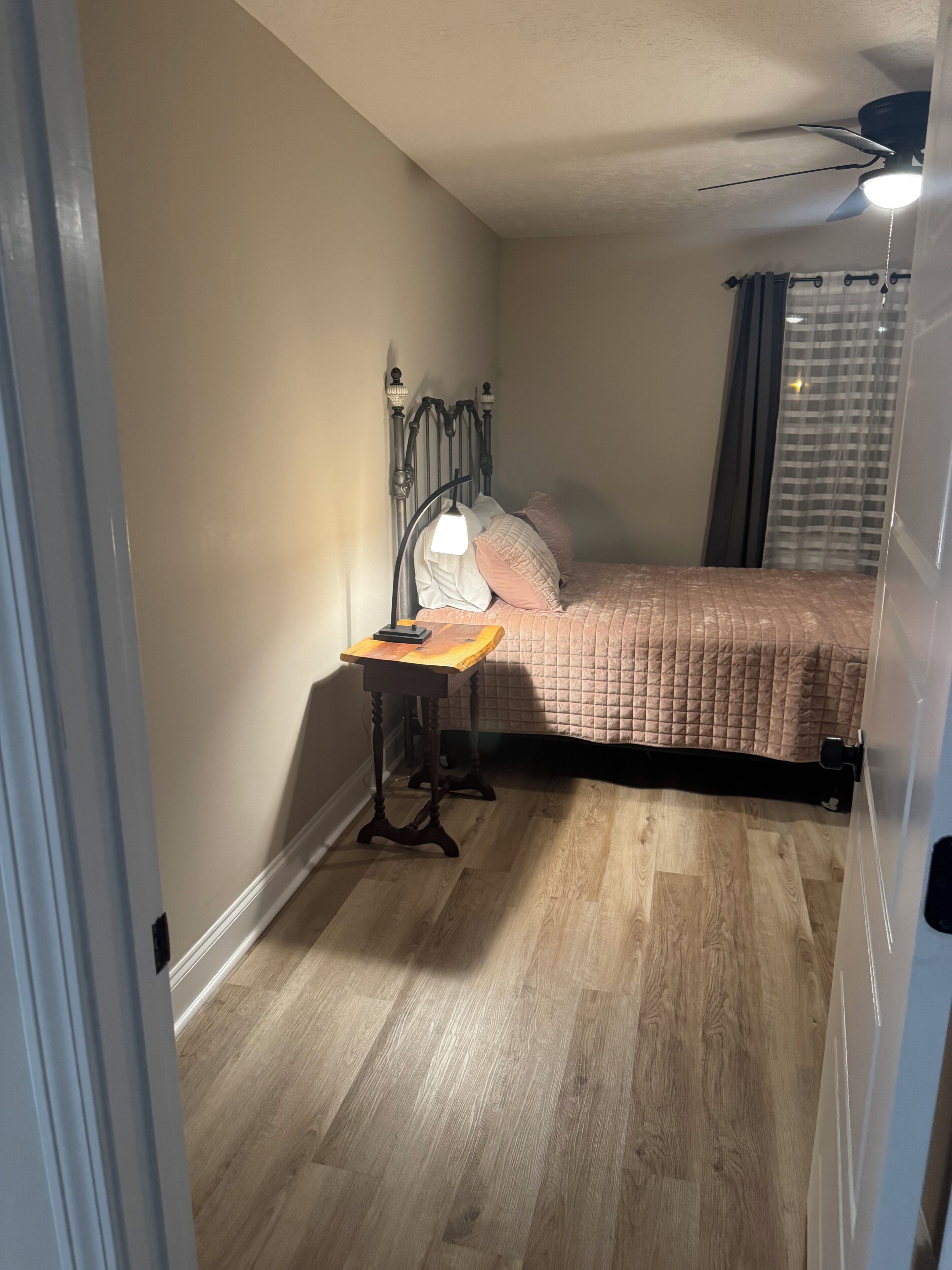 A bedroom with a bed, a bedside table with a lamp, a ceiling fan, and a window with curtains.