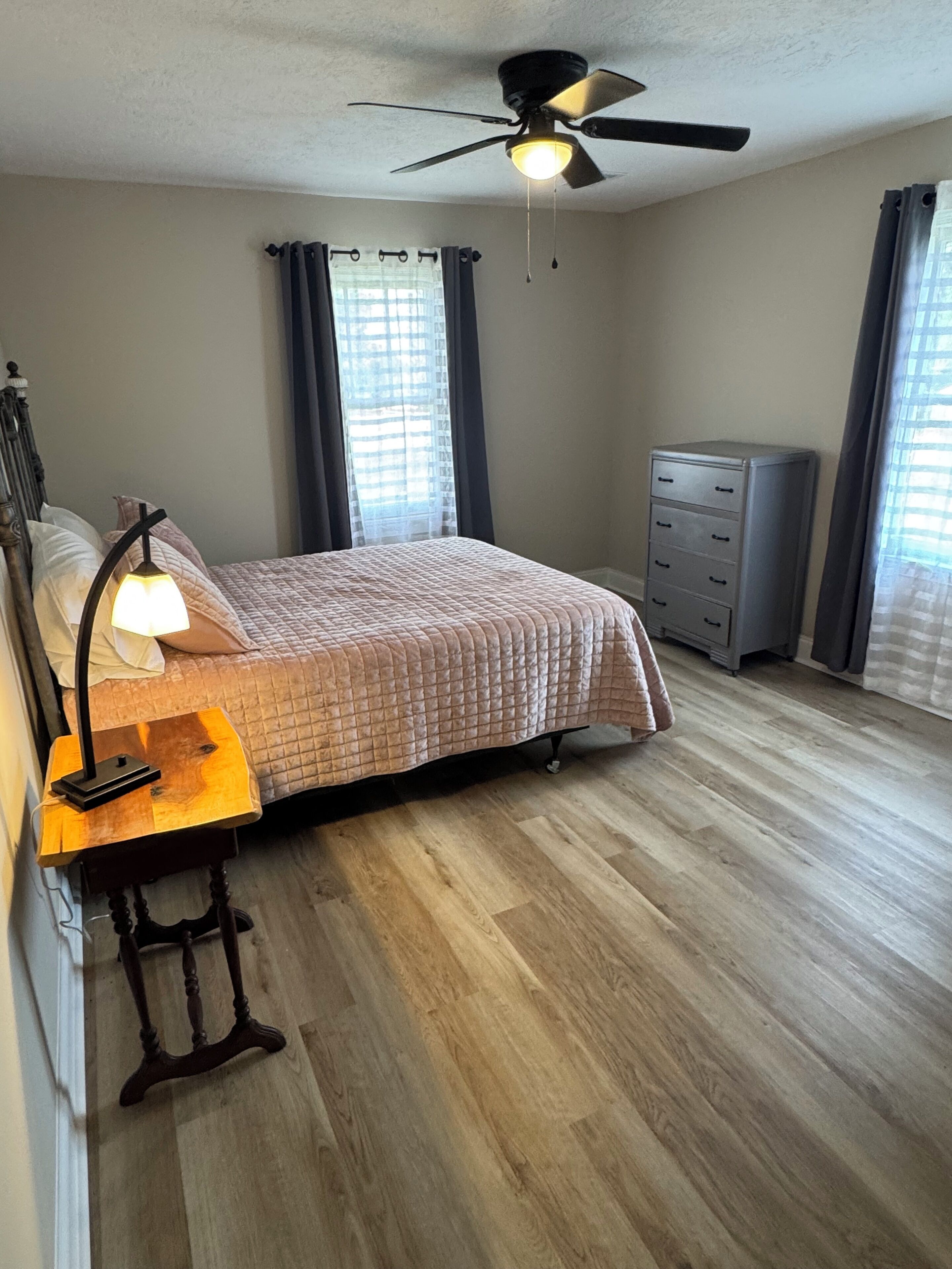 Une chambre à coucher comprenant un lit, un bureau avec une lampe, une commode, un ventilateur de plafond et une fenêtre avec des rideaux.