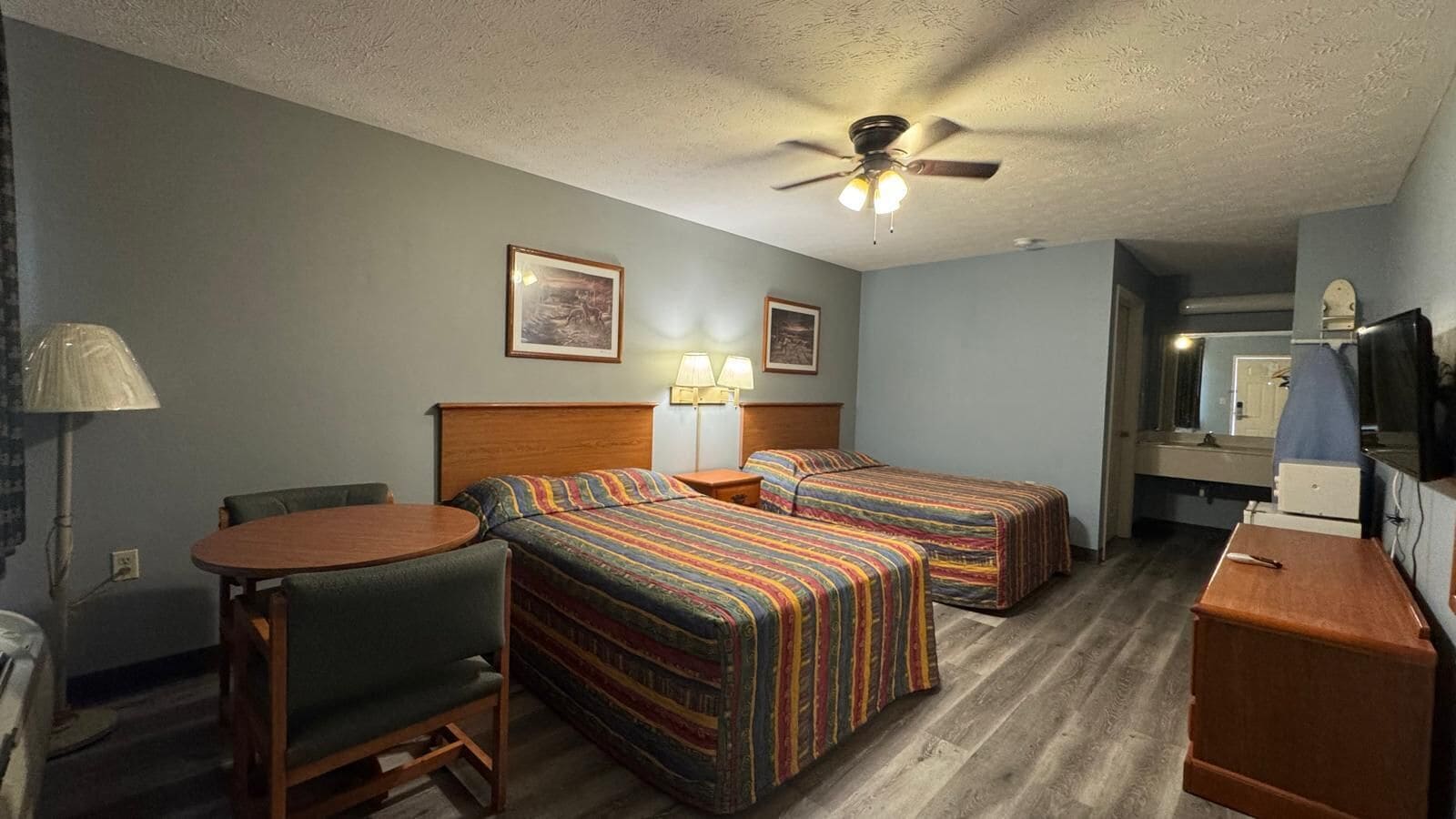 Habitación de hotel con dos camas, un escritorio, una silla, un ventilador de techo y un televisor.