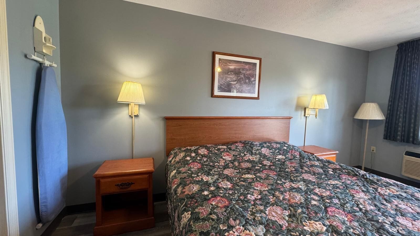 Habitación de hotel con cabecera de madera, colcha estampada, dos lámparas de noche, un cuadro en la pared y una cortina azul.
